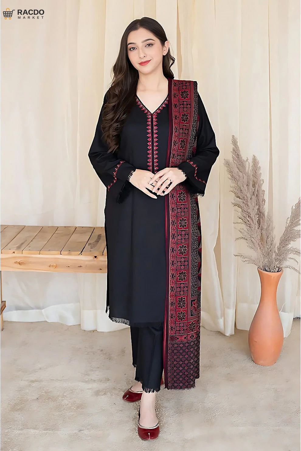Woman wearing a black and red traditional outfit with a patterned scarf, standing in a room with a wooden bench and vase.

